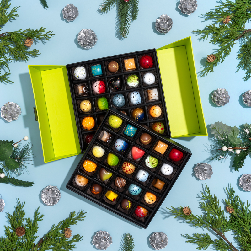 A stunning green tiered gift box featuring all 10 limited-edition holiday chocolates alongside 40 pieces from the signature collection. The gift box is displayed on a blue background with silver pinecones and evergreen branches.
